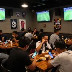 Penggemar merayakan selama pertandingan sepak bola langsung sambil memasang taruhan Judi Bola di smartphone mereka di bar olahraga.
